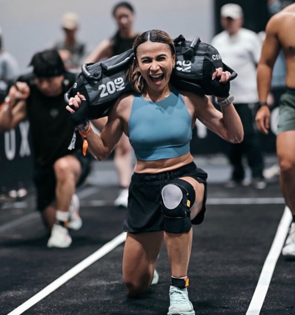 Dina laughing with a 20 kg sandbag on each shoulder mid-lunges