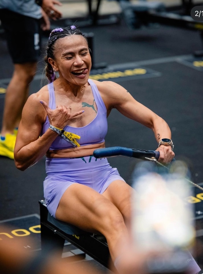 Dina laughing and flashing a shaka mid-pull on the RowErg in her lilac kit