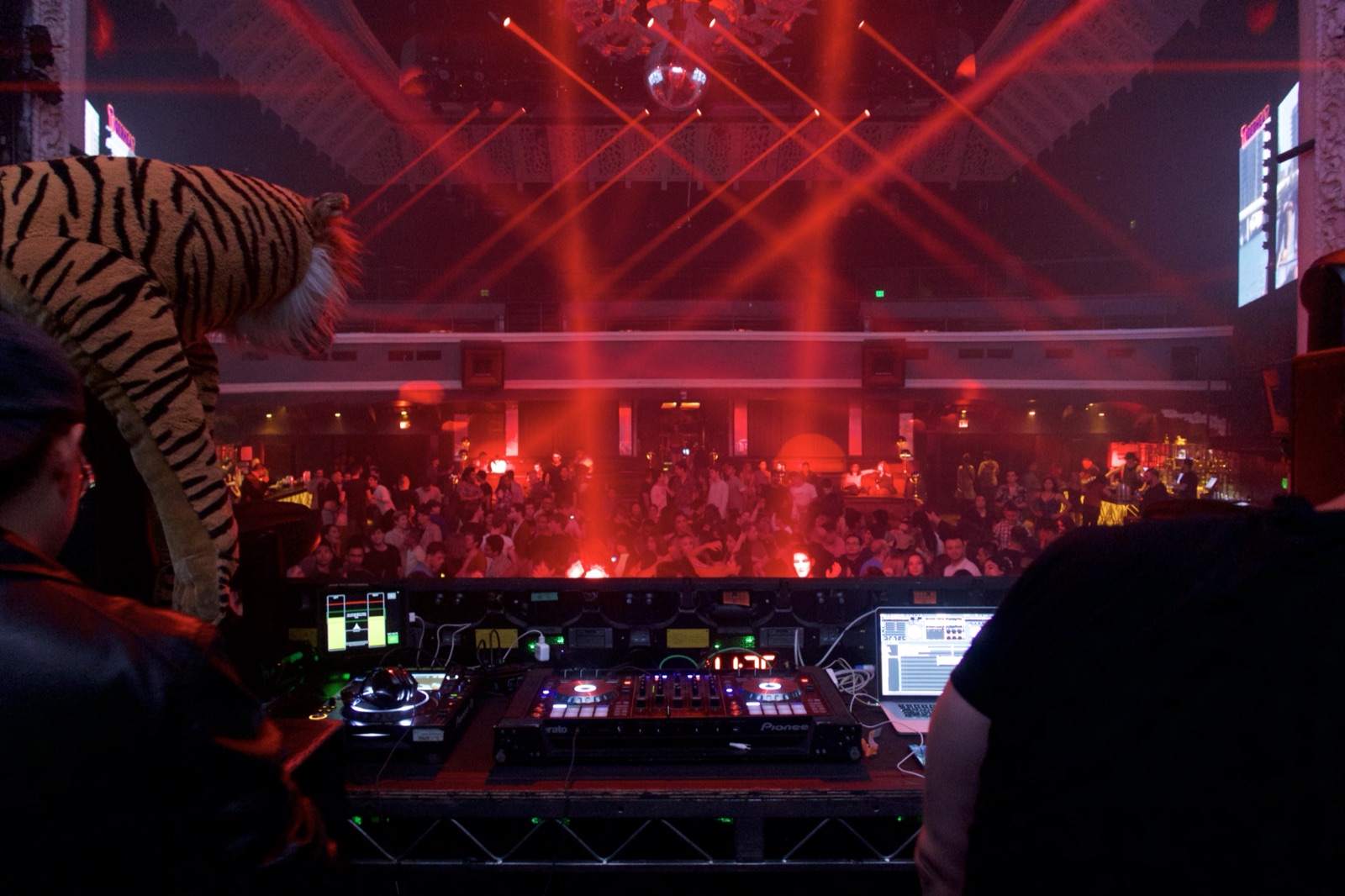A packed club dance floor seen from the DJ booth — red lasers, mirror ball, crowd with hands up